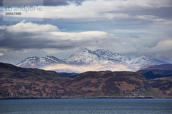 Hügel entlang der Küste und schneebedeckte Bergspitzen unter einem bewölkten Himmel; Argyll  Schottland'.