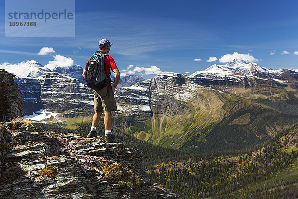 Männlicher Wanderer auf dem Gipfel eines Bergrückens mit Blick auf schneebedeckte Berge und das darunter liegende Waldtal mit blauem Himmel und Wolken; Waterton  Alberta  Kanada'.