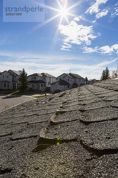 Nahaufnahme von gut abgenutzten abgerundeten Dachschindeln mit Häusern in der Nachbarschaft  Sonne  blauer Himmel und Wolken im Hintergrund; Calgary  Alberta  Kanada'.