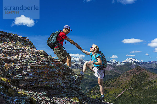 Männlicher Wanderer  der einer Wanderin auf einen felsigen Gipfel eines Bergrückens hilft  mit großartiger Aussicht und blauem Himmel und Wolken im Hintergrund; Waterton  Alberta  Kanada'.