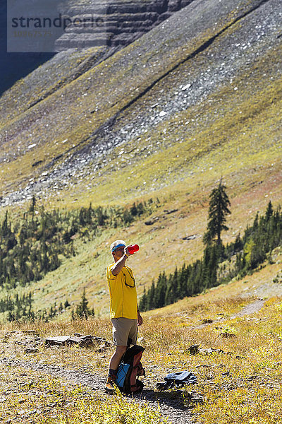 Männlicher Wanderer auf einem Wanderweg  der einen Schluck Wasser aus einer Wasserflasche nimmt  mit einem abfallenden Berghang im Hintergrund; Waterton  Alberta  Kanada'.