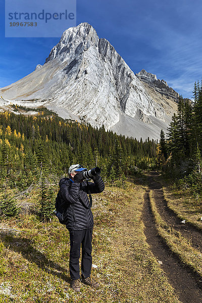 Männlicher Fotograf auf einem Bergpfad  der durch die Kamera schaut  mit Berggipfel und blauem Himmel im Hintergrund  Kananaskis Provincial Park; Alberta  Kanada'.