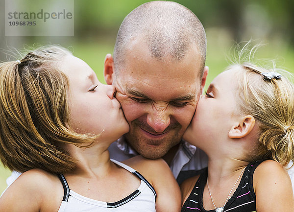 Ein Vater hält seine beiden Töchter  die ihn küssen  in einem Park; Edmonton  Alberta  Kanada'.