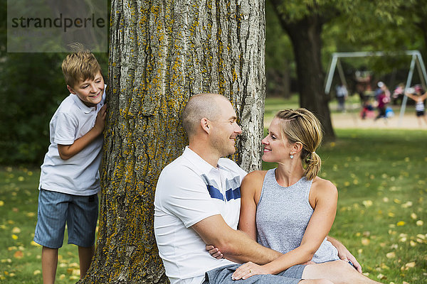 Ein Ehepaar  das während eines Familienausflugs in einem Park Zeit miteinander verbringt  während sein Sohn hinter einem Baum hervorschaut; Edmonton  Alberta  Kanada'.