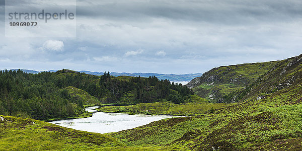 Ein Fluss  der in einen Teich fließt  umgeben von üppigem Gras und Wald in den Highlands; Schottland