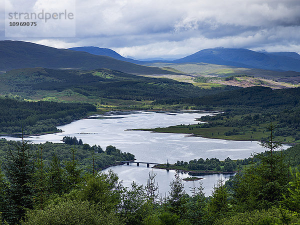 Fluss- und Waldlandschaft mit Bergen unter einem bewölkten Himmel; Schottland