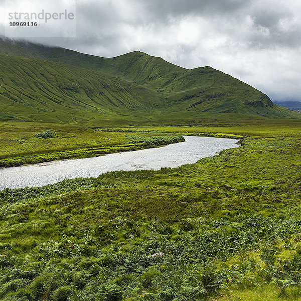 Berge und mit grünem Laub bedeckte Felder  durch die ein Fluss fließt  unter einem wolkenverhangenen Himmel in den Highlands; Schottland