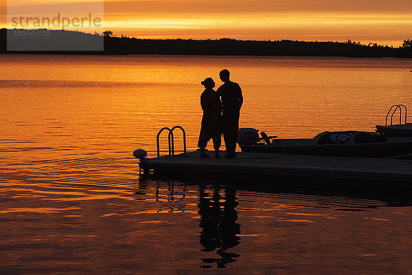 Silhouette eines Paares am Ende eines Stegs bei Sonnenuntergang mit orangefarbenem Licht am Himmel  das sich im ruhigen Wasser des Sees spiegelt; Ontario  Kanada'.