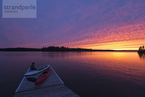 Eine junge Frau sitzt am Ende eines Stegs mit Kajaks und beobachtet den Sonnenuntergang über einem See; Ontario  Kanada'.