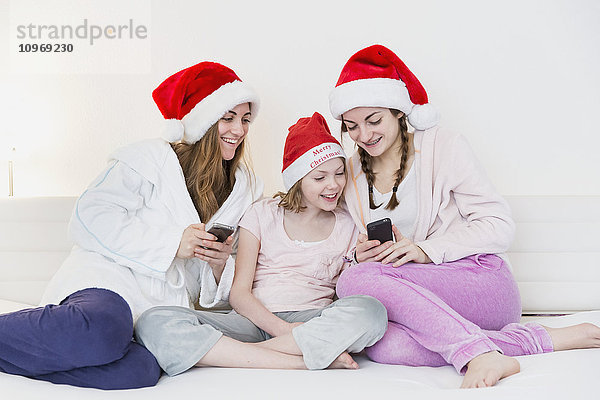 Drei Schwestern mit Weihnachtsmannmützen  Schlafanzügen und Gewändern  die zusammen auf einer Couch sitzen und lächelnd auf zwei Handys schauen; Bonn  Nordrhein Westfalen  Deutschland'.