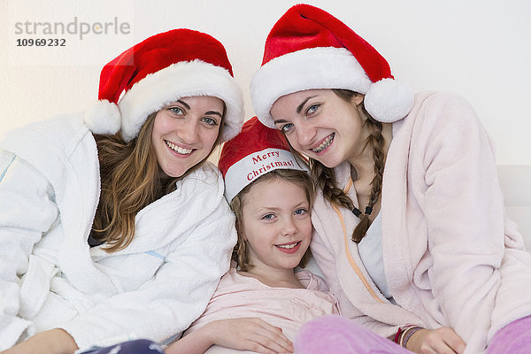 Drei Schwestern mit Weihnachtsmannmützen  Schlafanzügen und Gewändern  die zusammen auf einer Couch sitzen; Bonn  Nordrhein Westfalen  Deutschland