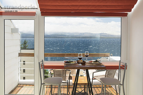 Ein Mittagstisch mit zwei Gläsern Rotwein ist vor dem Balkonfenster einer hellen Wohnung mit Blick auf einen See gedeckt; Bariloche  Argentinien'.