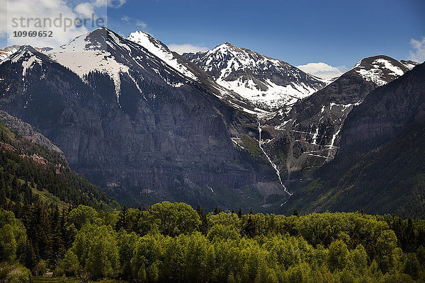 Schneebedeckte Berge von Telluride  Colorado  mit einem Wasserfall und einem Bach  der einen steilen Berghang hinunterstürzt  und sonnenbeschienenen Bäumen im Vordergrund; Colorado  Vereinigte Staaten von Amerika'.