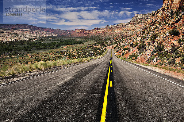 Utah Highway  der sich in die Ferne erstreckt  mit Felsklippen  die sich von der rechten Seite im Vordergrund bis zur Mitte und links im Hintergrund des Fotos erstrecken  mit blauem Himmel und einigen Wolken; Utah  Vereinigte Staaten von Amerika '