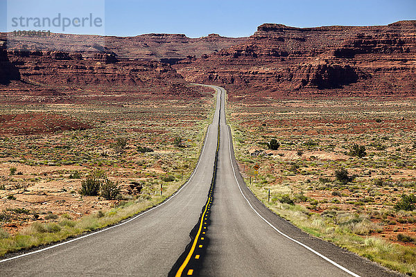 Utah Highway  der sich in die Ferne erstreckt  mit Felsklippen und blauem Himmel im Hintergrund und Wüstenvegetation auf beiden Seiten der Straße; Utah  Vereinigte Staaten von Amerika