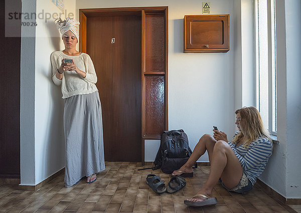 Zwei junge Frauen in ihrer Wohnung  die ihre Smartphones benutzen; Sagres  Portugal'.