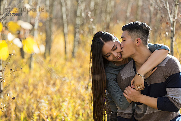 Ein junges asiatisches Paar genießt die gemeinsame Zeit im Freien in einem Park im Herbst und umarmt und küsst sich in der Wärme des Sonnenlichts; Edmonton  Alberta  Kanada'.