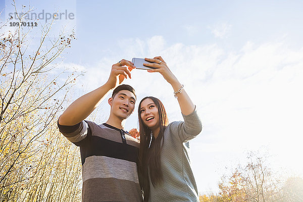 Ein junges asiatisches Paar genießt die gemeinsame Zeit in einem herbstlichen Park und macht Selbstporträts mit einem Smartphone in der späten Nachmittagssonne; Edmonton  Alberta  Kanada'.