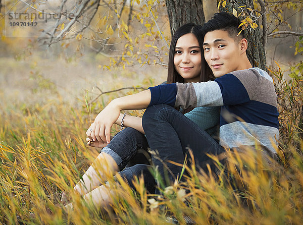 Ein junges asiatisches Paar genießt eine romantische Zeit im Freien in einem Park im Herbst unter einem Baum sitzend und für die Kamera posierend; Edmonton  Alberta  Kanada'.