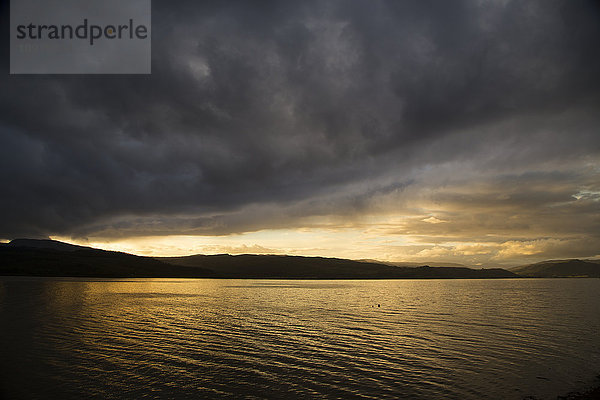 Sonnenlicht  das bei Sonnenuntergang durch die Wolken am Horizont leuchtet  mit einer Silhouette von Hügeln entlang der Küste; Strachur  Schottland'.