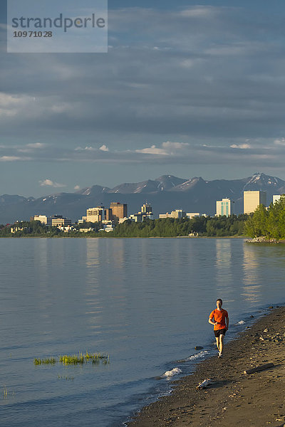 Mann beim Joggen am Strand neben dem Tony Knowles Coastal Trail während der Flut mit der Skyline der Stadt Anchorage  dem Knik Arm und den Chugach Mountains im Hintergrund; Anchorage  Alaska  Vereinigte Staaten von Amerika
