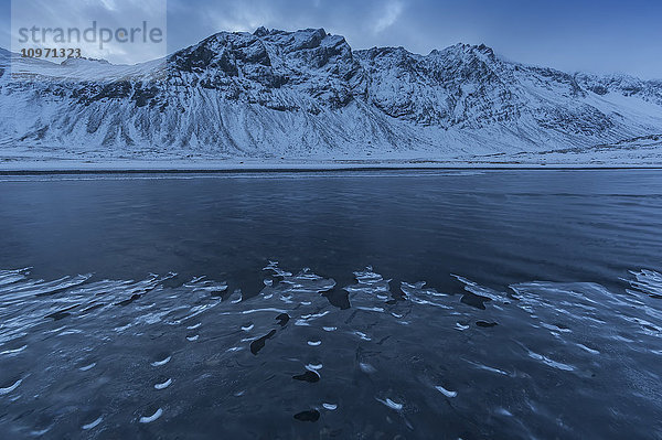 Schneebedeckte Landschaft entlang der Ostküste Islands  während ein Strom sanft vorbeifließt und sich Eis entlang des Ufers bildet; Island