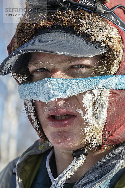 Porträt von Ben Harper mit Nasenmaske und frierend bei -40 Grad am Schild des Huslia-Kontrollpunkts während des Iditarod 2015