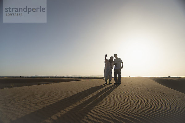 Silhouetten von Wüstenführer und Tourist bei Sonnenuntergang; Merzouga  Marokko'.