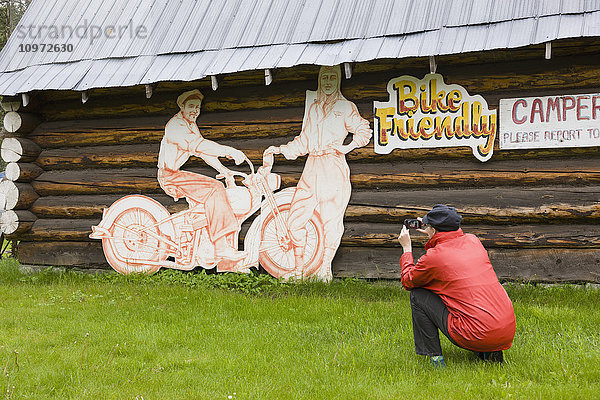 Eine Touristin fotografiert eine Blockhütte und ein Motorradschild an der Tetsa River Lodge  British Columbia  Kanada  Sommer