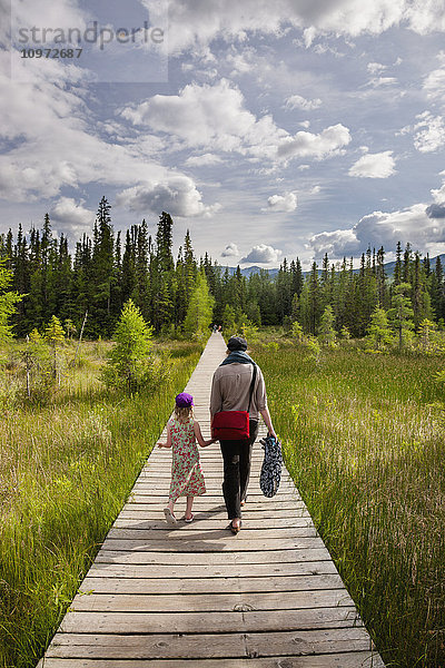 Eine Mutter und ihre Tochter spazieren auf einem Steg durch ein von immergrünen Bäumen umgebenes Feuchtgebiet  Liard River Hot Springs Provincial Park  British Columbia  Kanada  Sommer