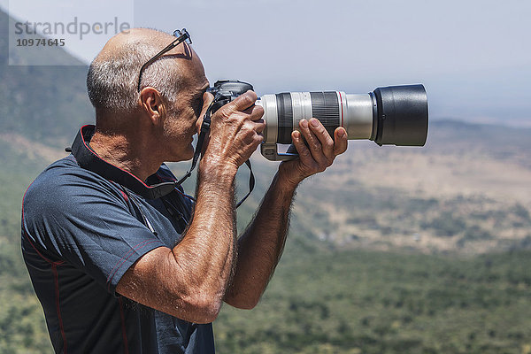 Ein glatzköpfiger Mann fotografiert mit einer digitalen Spiegelreflexkamera mit langem Objektiv einen Blick auf das Rift Valley im Sonnenschein; Tharaka Nithi  Kenia'.