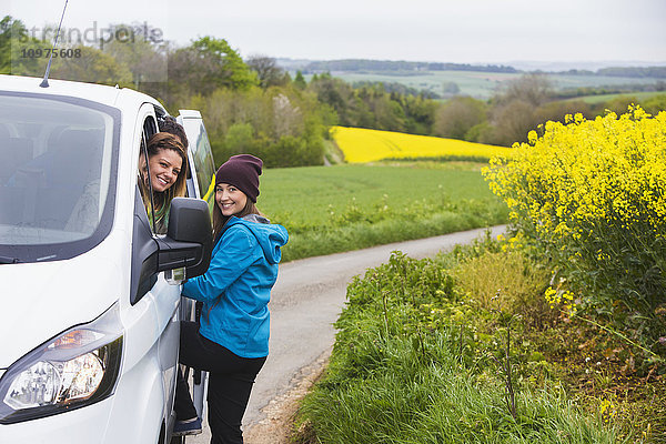 Zwei junge Frauen posieren vor ihrem Fahrzeug  während sie durch die englische Landschaft fahren  die mit leuchtend gelben Rapsblüten bedeckt ist; Cotswolds  England'.