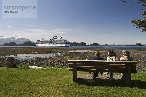 Touristen entspannen sich auf einer Bank entlang der Küste mit Blick auf das Kreuzfahrtschiff Celebrity Infinity  das in Sitka  Alaska  vor Anker liegt
