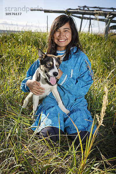 12 Jahre altes Athabascan-Mädchen in traditionellem Kuspuk  im Gras sitzend  mit Fischtrockengestell im Hintergrund  das einen Husky-Welpen hält  Nome  Alaska