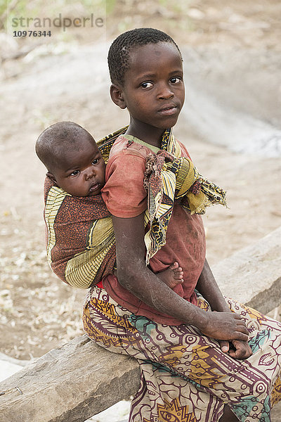 Junges Datoga-Mädchen trägt ihren kleinen Bruder auf dem Rücken  während sie auf einer Bank in der Nähe des Eyasi-Sees sitzt; Tansania'.