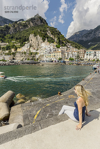 Eine junge Touristin posiert für ein Foto an der Strandpromenade mit der Stadt Amalfi im Hintergrund während eines Urlaubs in Italien; Amalfi  Provinz Salerno  Italien'.