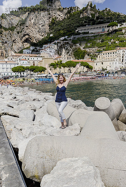 Eine junge Touristin posiert für ein Foto  aufgeregt für ihren Europa-Urlaub in Italien vor der Stadt Amalfi an der Amalfiküste mit dem Mittelmeer und Gebäuden im Hintergrund; Amalfi  Provinz Salerno  Italien'