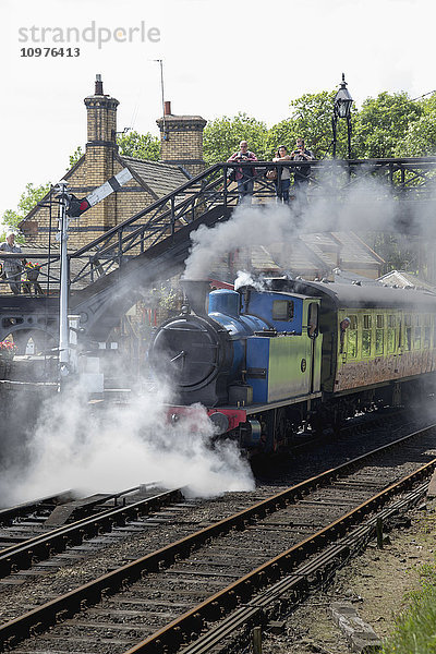 Dampf aus einem Personenzug  der die Gleise hinunterfährt  während Menschen von einem Steg über den Gleisen zusehen; Cumbria  England'.