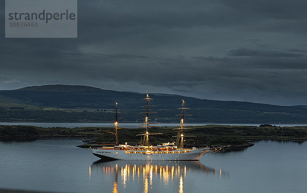Ein großes weißes Boot mit beleuchteten Masten auf dem ruhigen Wasser entlang der Küste in der Abenddämmerung; Tobermory  Isle of Mull  Argyll und Bute  Schottland'.
