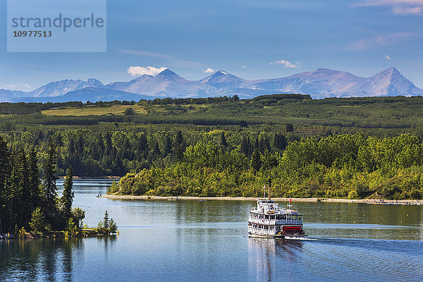 Schaufelradboot auf einem See mit baumgesäumter Uferlinie  Hügeln und Bergen im Hintergrund und blauem Himmel und Wolken; Calgary  Alberta  Kanada