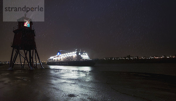 Licht  das von einem Leuchtturm an der Küste ausgeht  während ein beleuchtetes Kreuzfahrtschiff vorbeifährt; South Shields  Tyne and Wear  England'.