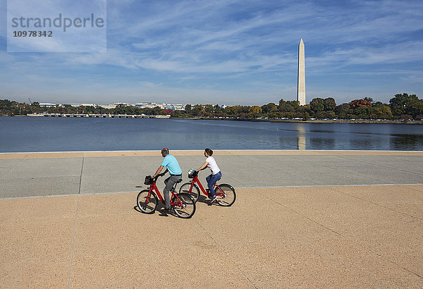 Besucher fahren mit Bikeshare-Fahrrädern entlang des Tidal Basin direkt vor dem Thomas Jefferson Memorial mit dem Washington Monument im Hintergrund; Washington  District of Columbia  Vereinigte Staaten von Amerika'.