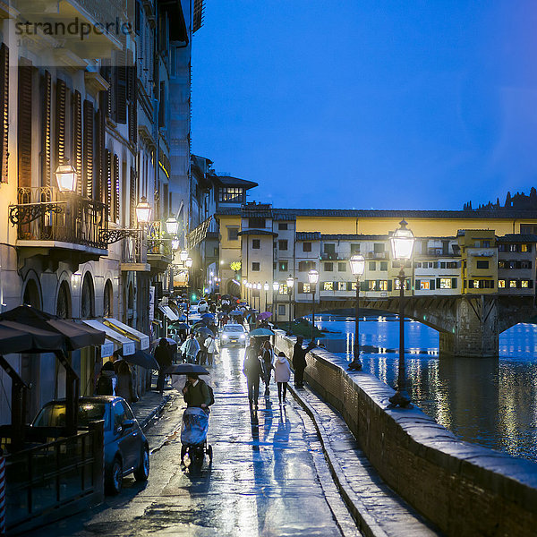 Fußgänger auf einer nassen Promenade in der Abenddämmerung entlang des Arno mit Blick auf die Ponte Vecchio; Florenz  Italien'.