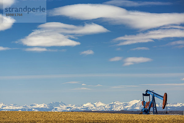 Pumpenheber in einem Stoppelfeld mit schneebedeckten Bergen im Hintergrund mit blauem Himmel und Wolken; Alberta  Kanada'.