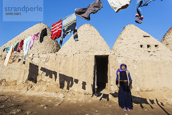 Eine Frau steht vor der Tür ihrer primitiven Behausung im Schatten der auf der Wäscheleine hängenden Kleidung; Harran  Türkei