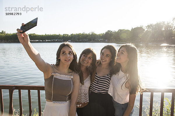 Vier Freunde stehen im Sonnenlicht vor dem Wasser und nehmen Selfie mit dem Smartphone.