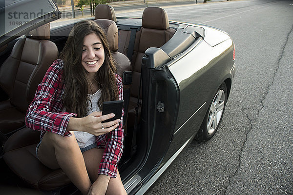 Lächelnde junge Frau sitzt im Cabrio und schaut auf das Smartphone.