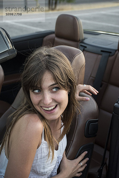Lächelnde junge Frau sitzt im Cabrio und zieht lustige Gesichter.