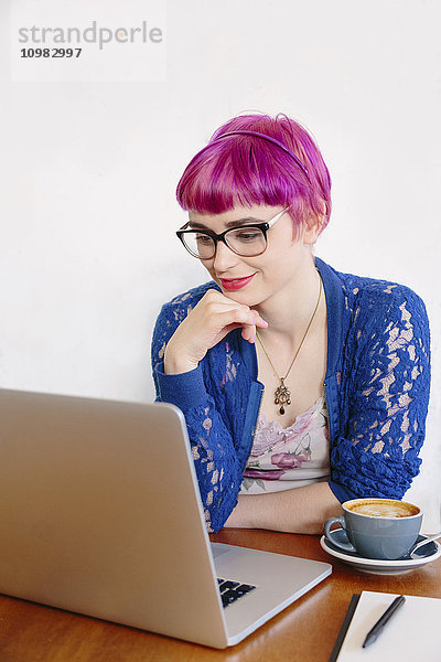 Porträt einer lächelnden jungen Frau mit gefärbten Haaren  die in einem Café sitzt und auf den Laptop schaut.