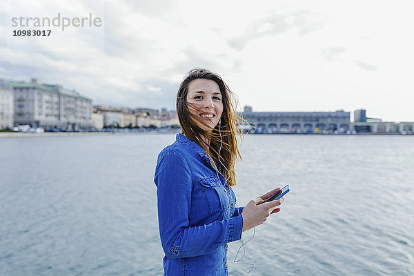 Italien  Triest  lächelnde junge Frau beim Musikhören am Wasser